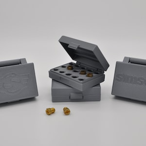 Könnte beinhalten: Drei graue Plastikboxen mit Deckeln, zwei geschlossen und eine offen. Die offene Box enthält einen Satz kleiner, goldfarbener Metallteile. Die Boxen haben die Worte "Smilsen" und "S" mit Flügeln aufgedruckt.