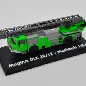 Op de afbeelding: Een groen en grijs model brandweerwagen met een uitschuifbare ladder, gemonteerd op een zwarte basis. Het model is gemarkeerd met "Magirus DLK 23/12 - Maßstab 1:87". De brandweerwagen heeft gedetailleerde ontwerpelementen en is een miniatuur replica.