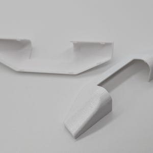 May include: Two white plastic pieces, possibly for a model car or other project. One piece is a curved, rounded shape, while the other is a more angular, flat piece.