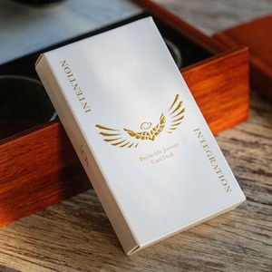 Puede incluir: Caja blanca con letras doradas y un símbolo de alas doradas. La caja dice "Integración Intención Viaje Psicodélico Juego de Cartas".