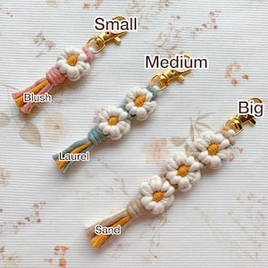 May include: Three crocheted daisy flower keychains in different sizes: small, medium, and large. The small keychain is blush pink, the medium keychain is teal, and the large keychain is cream with a yellow center. The keychains are labeled "Small", "Medium", "Big", "Blush", "Laurel", and "Sand".