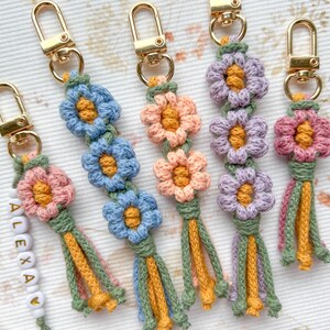 May include: Five crocheted flower keychain charms with gold key rings. The charms are made with pink, purple, blue, and yellow yarn. The charms have a tassel at the bottom.