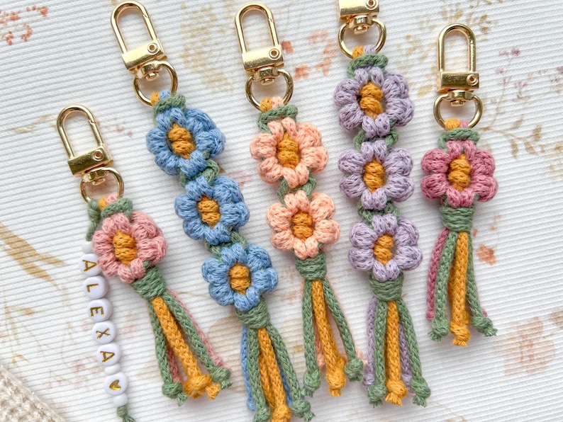 May include: Five crocheted flower keychain charms with gold key rings. The charms are made with pink, purple, blue, and yellow yarn. The charms have a tassel at the bottom.