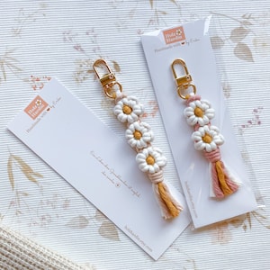 May include: Two handmade daisy keychain charms with pink and yellow tassels. The charms are made with white and yellow string and have a gold key ring. The charms are displayed on white cards with the text "Holz Hardin Handmade with love by Erika" and the website address "holzhardin.etsy.com".