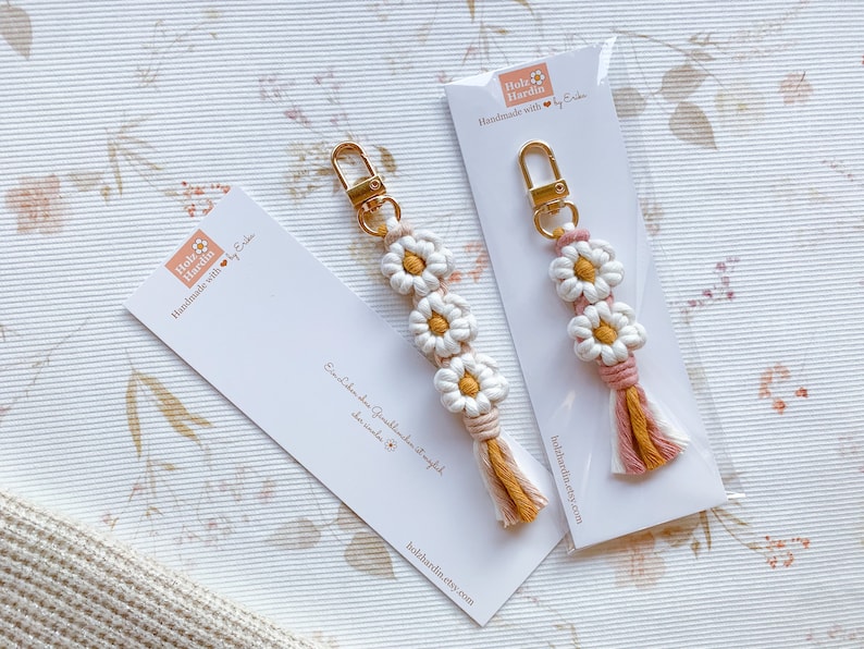 May include: Two handmade daisy keychain charms with pink and yellow tassels. The charms are made with white and yellow string and have a gold key ring. The charms are displayed on white cards with the text "Holz Hardin Handmade with love by Erika" and the website address "holzhardin.etsy.com".