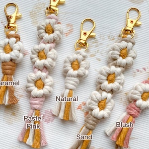 May include: Five handmade macrame keychains with daisy-shaped designs in various colors, including caramel, pastel pink, natural, sand, and blush. Each keychain features a gold metal key ring.