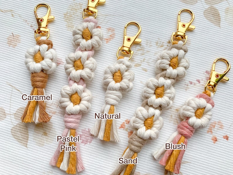 May include: Five handmade macrame keychains with daisy-shaped designs in various colors, including caramel, pastel pink, natural, sand, and blush. Each keychain features a gold metal key ring.