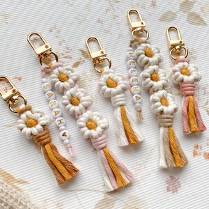 May include: Five colorful keychains with daisy-shaped charms and beaded tassels. The keychains are made of macrame and feature the names "Sarah", "Helena", and "Ava" in white beads.