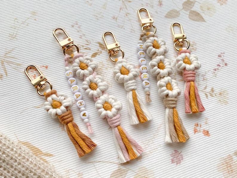May include: Five colorful keychains with daisy-shaped charms and beaded tassels. The keychains are made of macrame and feature the names "Sarah", "Helena", and "Ava" in white beads.