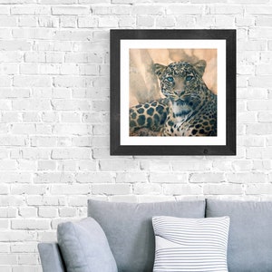Op de afbeelding: Een ingelijste print van een luipaard met blauwe ogen, liggend en rechtstreeks naar de kijker kijkend. De achtergrond is een aquarel in bruin en beige tinten.