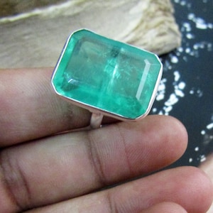 May include: A large, rectangular emerald-cut gemstone ring. The ring features a vibrant, transparent green stone set in a silver-coloured metal band. The gemstone has a faceted surface, reflecting light. The ring is a statement piece of jewellery.