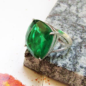 Peut inclure: Une bague en argent ornée d'une grosse pierre précieuse vert émeraude. La pierre, de taille coussin, est sertie dans un anneau en argent. La bague est présentée sur une surface grise texturée avec un fond blanc et orange.