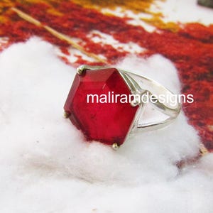 Op de afbeelding: Een zilveren ring met een grote, facetgeslepen, rode edelsteen in een zeshoekige vorm. De tekst "maliramdesigns" is op de edelsteen gedrukt. De ring is geplaatst tegen een witte en kleurrijke achtergrond.