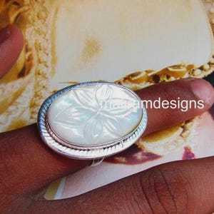 Könnte beinhalten: Ovale Silberring mit einem geschnitzten weißen Blumendesign. Der Ring hat einen strukturierten Rand und sitzt auf einem Finger. Der Ring ist ein Schmuckstück.