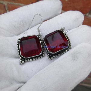 May include: Silver earrings featuring large, square, deep red gemstones. The gemstones are set in a silver frame with a beaded border. The earrings have a simple hook closure.