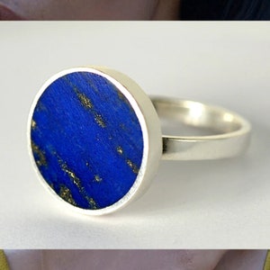 Könnte beinhalten: Ein silberner Ring mit einem großen, runden, tiefblauen Stein mit Goldeinschlüssen. Der Stein ist in einem polierten Silberband gefasst. Der Ring hat ein minimalistisches Design, geeignet für den Alltag oder besondere Anlässe. Ein Schmuckstück.