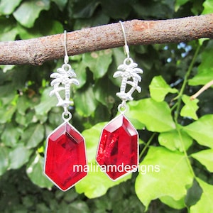 May include: A pair of silver-toned earrings with a palm tree design at the top and a large, faceted, red gemstone pendant. The earrings are hanging from a brown branch against a backdrop of green foliage. The text "maliramdesigns" is visible.