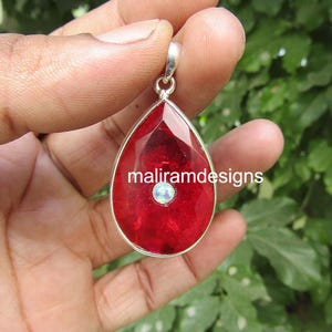 May include: A teardrop-shaped pendant with a vibrant red gemstone, accented by a small, iridescent stone at the center. The pendant is set in a silver-colored metal frame. The text "maliramdesigns" is visible on the pendant.