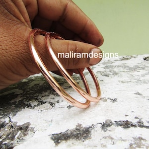 Op de afbeelding: Drie koperkleurige armbanden worden in een hand gehouden. De armbanden zijn glad en glanzend en reflecteren licht. De tekst "maliramdesigns" is zichtbaar op de hand. De achtergrond is een gevlekt grijs en wit oppervlak.