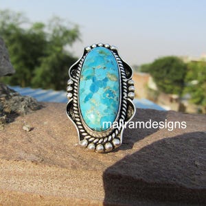May include: A silver ring with a large, oval turquoise stone. The stone is a vibrant blue with brown and gold veining. The ring has a decorative silver setting with a beaded border and the text "malramdesigns".