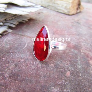 May include: A silver ring featuring a large, teardrop-shaped, faceted red gemstone. The gemstone is set in a polished silver band. The ring is displayed on a textured, reddish-brown surface. The text "maliramdesigns" is visible on the gemstone.