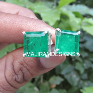 May include: A pair of square-cut emerald earrings with a silver setting. The vibrant green gemstones are set in a four-prong design. The earrings are held in a hand, with the text "MALIRAMDESIGNS" visible.