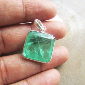 May include: A square, emerald-green gemstone pendant with a silver bail. The gemstone is faceted, reflecting light. The pendant is held in a hand.