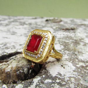 May include: A gold-colored ring with a rectangular, deep red gemstone at its center. The gemstone is surrounded by a border of small, clear stones. The ring has a decorative, openwork design and the text "iramdesigns" is visible.