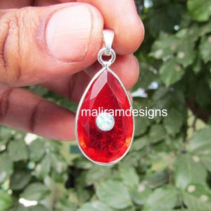May include: A teardrop-shaped pendant with a faceted red gemstone and a small, round, white gemstone at the center. The pendant has a silver-colored metal frame and a bail. The text "maliramdesigns" is printed on the pendant.