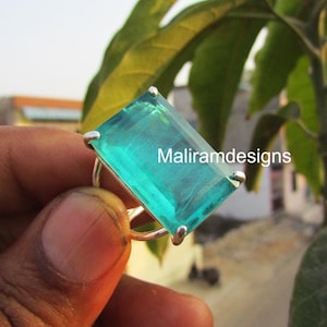 May include: A silver ring with a large, rectangular, teal gemstone. The gemstone is held in place by four silver prongs. The ring is shown against a blurred backdrop of green leaves. The text "Maliramdesigns" is visible on the gemstone.
