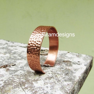 Op de afbeelding: Een koperen manchetarmband met een gehamerde textuur. De open armband heeft een gepolijste afwerking. De tekst "galiramdesigns" is zichtbaar aan de rechterkant. De armband wordt gepresenteerd op een grijs, getextureerd oppervlak.