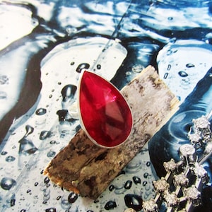 Puede incluir: Un anillo de plata con una gran piedra preciosa roja facetada en forma de lágrima. El anillo está colocado sobre un trozo de madera sobre un fondo de gotas de agua y un collar de plata.