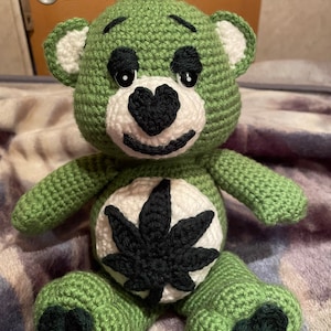 Può includere: Un orsacchiotto all'uncinetto verde fatto a mano con un naso nero a forma di cuore e una foglia di marijuana nera sulla pancia. L'orso ha accenti bianchi sulle orecchie e un piccolo ciuffo di filo nero sulla testa. L'orso è seduto su una coperta viola e grigia.