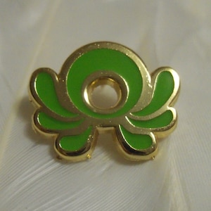 May include: A gold-colored pin with a green enamel design. The design is a stylized swirl with a hole in the center.