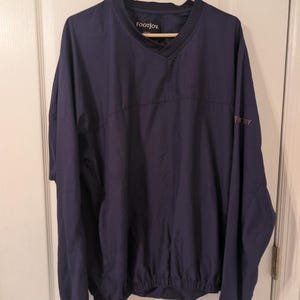 May include: A navy blue long-sleeve pullover windbreaker featuring a V-neck collar and an elasticized hem. The garment displays the FootJoy brand name on the interior neck label and a small embroidered FootJoy logo on the left sleeve.