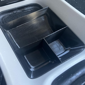 Könnte beinhalten: Ein schwarzer Auto-Organizer aus Kunststoff mit vier Fächern. Der Organizer ist so konzipiert, dass er in die Mittelkonsole eines Autos passt.