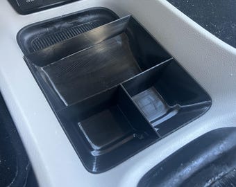 May include: A black plastic car organizer with four compartments. The organizer is designed to fit in a car's center console.