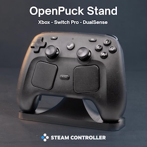 May include: A black OpenPuck Stand for Xbox, Switch Pro, and DualSense controllers, holding a black Steam Controller. The text OpenPuck Stand, Xbox - Switch Pro - DualSense, and STEAM CONTROLLER are visible on the image with a logo.
