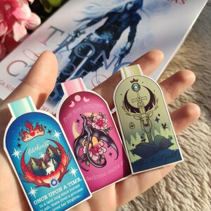 May include: Three colourful bookmarks with illustrations and text. The first bookmark is blue with a white wolf, a crown, and the text "filcheurt" and "ONCE UPON A TIME, In a land long since burned to ash, there lived a princess who loved her kingdom..." The second bookmark is pink with a black dragon, flowers, and the text "from now until the darkness claims us". The third bookmark is green with a deer, a sword, and the text "The law is not gold".
