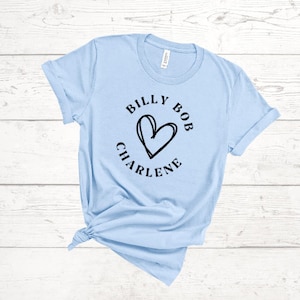 May include: Light blue t-shirt with the text "BILLY BOB CHARLENE" encircling a black heart design. The shirt is made of a soft material and has short sleeves. The shirt is knotted at the bottom.
