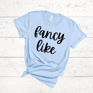 May include: A light blue t-shirt with the words "fancy like" in black script. The shirt is made of a soft material and has short sleeves. The shirt is tied at the bottom.