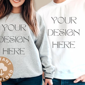 Op de afbeelding: Twee sweatshirts, &eacute;&eacute;n grijs en &eacute;&eacute;n wit, met de tekst "YOUR DESIGN HERE" gedrukt op de voorkant. Het grijze sweatshirt heeft een kleine bruine cirkel met een wolk en een downloadpijl-icoon.
