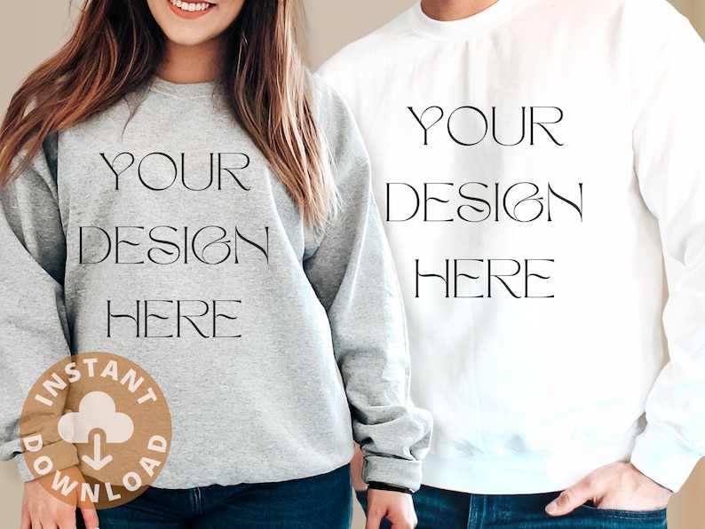 Op de afbeelding: Twee sweatshirts, &eacute;&eacute;n grijs en &eacute;&eacute;n wit, met de tekst "YOUR DESIGN HERE" gedrukt op de voorkant. Het grijze sweatshirt heeft een kleine bruine cirkel met een wolk en een downloadpijl-icoon.