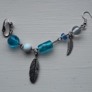 May include: A unique earring featuring a silver-tone clip-on and hook design. The earring is adorned with turquoise and white beads, connected by a delicate chain. Two silver feather charms dangle from the chain, adding a touch of whimsy.