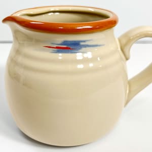 May include: A cream-colored ceramic creamer with a brown rim and a blue and red design. The creamer has a rounded body and a handle.