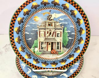 Puede incluir: Un plato redondo de cerámica con una ilustración de una casa encantada con un fantasma, murciélagos y calabazas. El borde tiene un patrón de cuadros negros y naranjas con murciélagos y lunas amarillas. El centro muestra una casa con el texto Welcome y Notice.