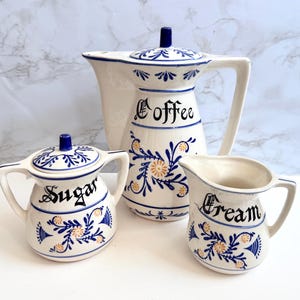 May include: A ceramic coffee service set featuring a coffee pot, a sugar bowl with a lid, and a cream pitcher. Each piece is white with blue floral patterns, yellow accents, and black script text reading Coffee, Sugar, and Cream respectively.