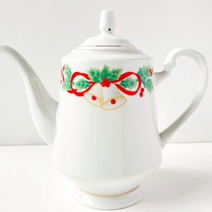 May include: White ceramic teapot with a Christmas design featuring red bows, green holly leaves, and gold bells.