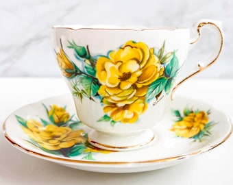 May include: A white teacup and saucer set featuring a vibrant yellow rose floral design with green leaves. The cup and saucer have gold trim. The teacup has a handle and sits on a small pedestal base. The saucer is scalloped.