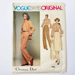 May include: A vintage Vogue Paris Original sewing pattern envelope, number 1608, featuring Christian Dior designs. The cover displays a photograph and illustrations of a tan jacket, trousers, a striped blouse, and a skirt, with text in blue and red.